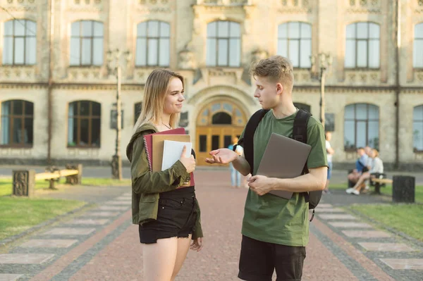 Пара молодих привабливих студентів розмовляють на відкритому повітрі в парку біля будівлі університету, тримаючи ноутбук, книги та блокноти. Двоє студентів коледжу розмовляють біля кампусу . — стокове фото