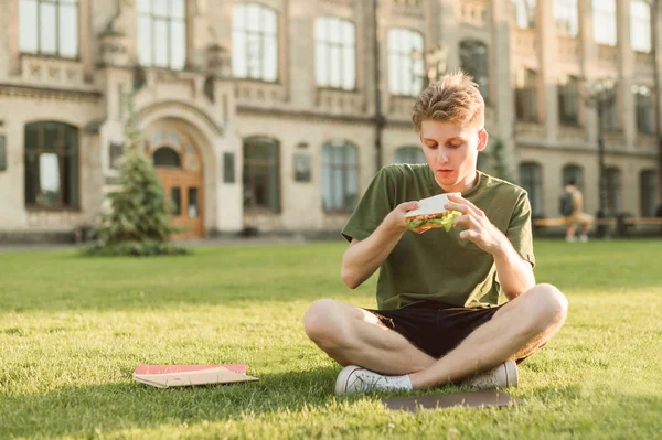 Un bel ragazzo in abiti casual seduto su un prato sullo sfondo di un edificio universitario con un panino in mano e andare a mangiare. Ritratto di uno studente affamato che mangia un panino in pausa . — Foto Stock