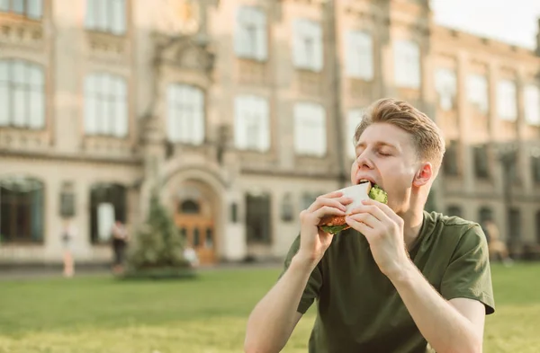 Retrato de cerca de un joven sentado en el fondo del edificio, mordiendo un sándwich con los ojos cerrados.Estudiante hambriento come una comida apetitosa del hombre en el fondo de un edificio de la universidad — Foto de Stock