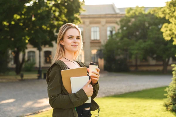 Feche a foto de uma menina alegre que anda na rua perto da escola que mantém livros e café. Sorrindo menina da faculdade em casaco verde andando perto do campus da faculdade com tirar café e notebook . — Fotografia de Stock