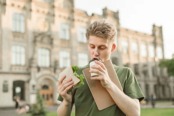 Busy boy eats a fast-food university campus, he does not have time to.Hungry young boy eats a sandwich and drinks coffee from a cup on the back of the university building, wears a green t-shirt — Stock Photo, Image