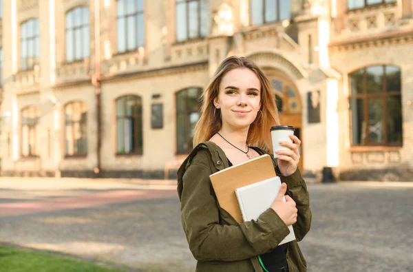 Portré fiatal csinos vidám nő visel zöld kabát gazdaság könyveket és kávéscsésze mellett a főiskolai campus. Pozitív női diák az utcán egyetemi udvarban, notebookokkal és kávéval. — Stock Fotó