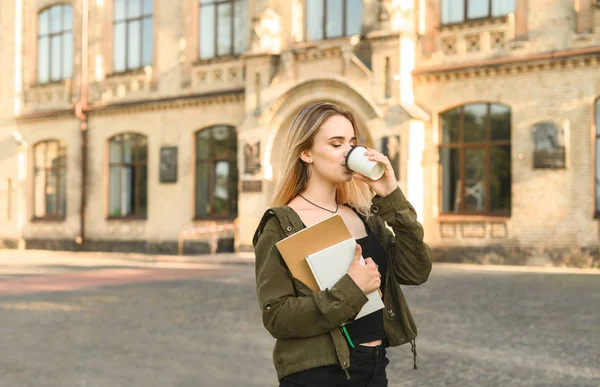 Student drinking coffee concept. Happy young female student enjoying take away coffee holding notebooks near college campus. Positive relaxed student drinks coffee from paper cup near the university.