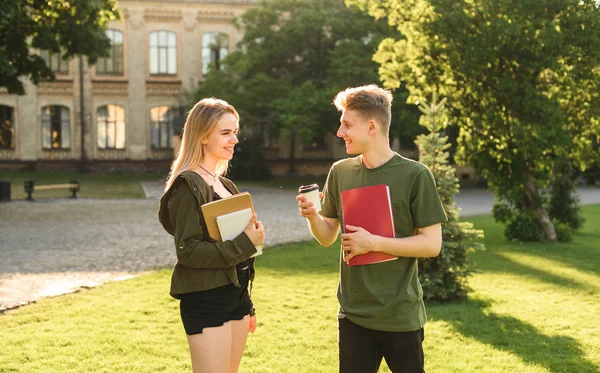 Двоє привабливих студентів, жінка і чоловік, проводять розмову в парку біля університету, тримаючи ноутбуки і каву. Молоді студенти спілкуються парами біля коледжу . — стокове фото