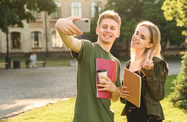 Jovens amigos do sexo feminino e masculino no parque posando para selfie na câmera do smartphone, estudantes milenares tirando fotos no celular segurando cadernos enquanto fazem uma pausa para o café no campus da faculdade . — Fotografia de Stock