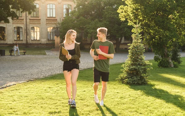 Twee studenten, vrouwelijke en mannelijke, vrolijk lopen in het Universiteits Park houden hun notebooks. Paar studenten terloops lopen in College Garden met een gesprek. Succesvol onderwijsconcept. — Stockfoto
