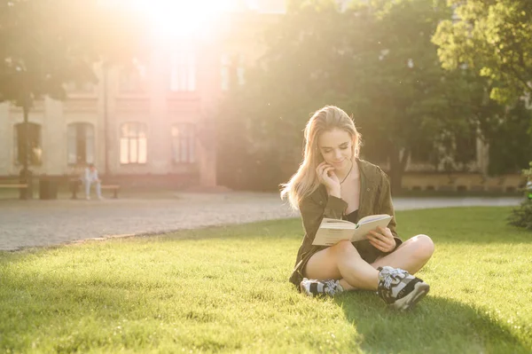 Linda estudiante joven sentada en la hierba y leyendo el libro en el parque universitario. Estudiante encantada leyendo el libro cerca del edificio de la universidad en el patio . —  Fotos de Stock