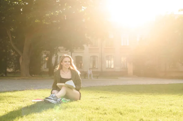 Jovem menina estudante bonita sentado na grama parque campus sob a luz do sol com livros. Estudante sorridente estudando e se preparando para o exame na universidade. Conceito de ensino universitário . — Fotografia de Stock
