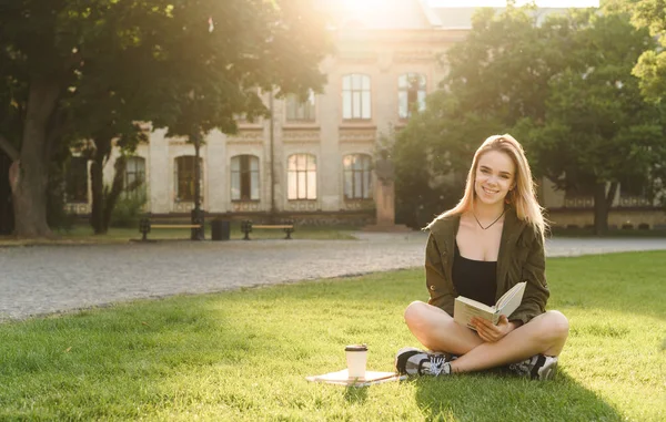 大学の公園に座って宿題をしながらカメラを見て幸せな学生。大学の庭の芝生に座って、授業の準備をしている本を持つ笑顔の若い学生. — ストック写真