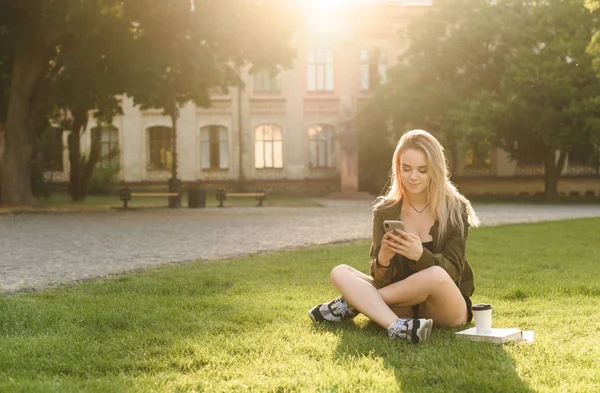 大学の庭の芝生の上に座りながら、スマートフォンを使った若い大学生の女の子。彼女の携帯電話にテキストメッセージを公園に座って大学の建物の近くに肯定的な女子学生. — ストック写真