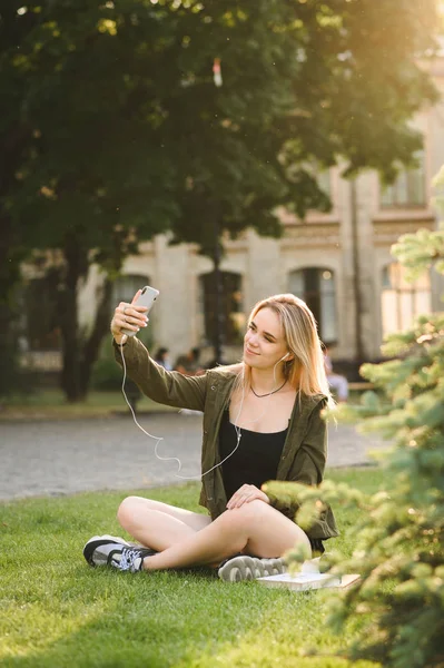 Счастливая студентка-подросток в чате смотрит на смартфон, посылает видео-сообщение, делает селфи позирует, урок на улице, сидит на лужайке зеленого колледжа. Вертикальный выстрел . — стоковое фото