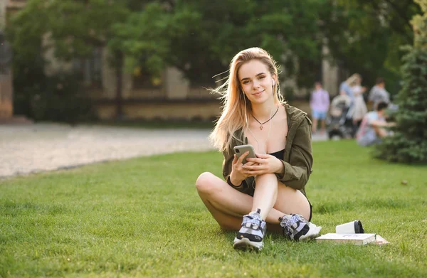 Güneşli bir günde akıllı telefonunu kullanarak çimlerin üzerinde oturan bir kız üniversite öğrencisinin portresi. Kafkas lise öğrencisi kız üniversite parkı telefonda onu kulaklık ile müzik dinliyor. — Stok fotoğraf
