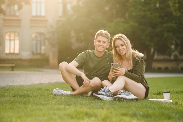 Молода весела кавказька мила пара студентів коледжу, які сидять на траві біля унікального кампусу, слухаючи музику з навушниками. Двоє милих студентів використовують смартфон та навушники у парку біля коледжу . — стокове фото