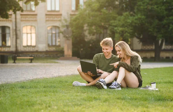 Twee jonge college studenten kijken naar grappige video op de laptop, voorbereiden voor het examen met computer zittend op het gazon. Paar studenten in het Universiteits Park met behulp van laptop, educatie en leren. — Stockfoto
