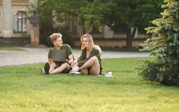 Um casal atraente de estudantes caucasianos sentados no relvado perto do campus. Bonito estudante do sexo masculino segurando o laptop, olhando para a menina estudante, tendo uma conversa no parque universitário . — Fotografia de Stock