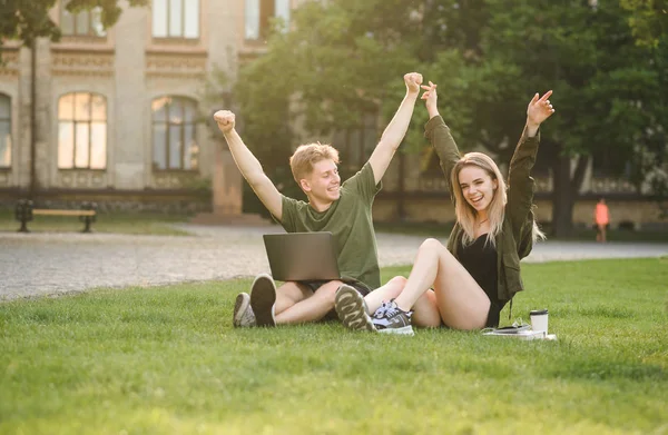 Couple of successful young university students raising hands up into two sides feeling proud and happy sitting on lawn. Two cheerful students holding hands up feeling like winners near college campus.