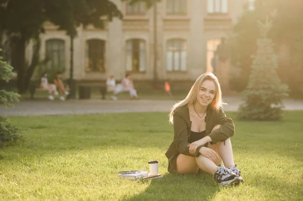 Jovem estudante atraente do sexo feminino com um sorriso largo sentado no gramado no quintal da faculdade com café e livros. Sorrindo menina estudante alegre ficar no parque universitário, relaxante com tomar café . — Fotografia de Stock