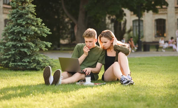 Figyelmes tini diákok pár kereső tartalmat, kommentálva laptop ül a füvön az egyetemi parkban. Koncentrált diákok látszó-on laptop képernyő, vizsgázó információ-ban kollégium kert. — Stock Fotó