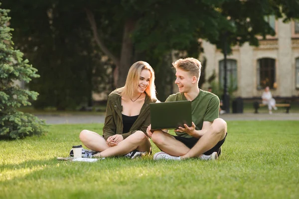 Happy college students communicating, talking, using laptop on campus lawn. Young cheerful caucasian couple of students watching videos on computer, looking at the screen in the university park.