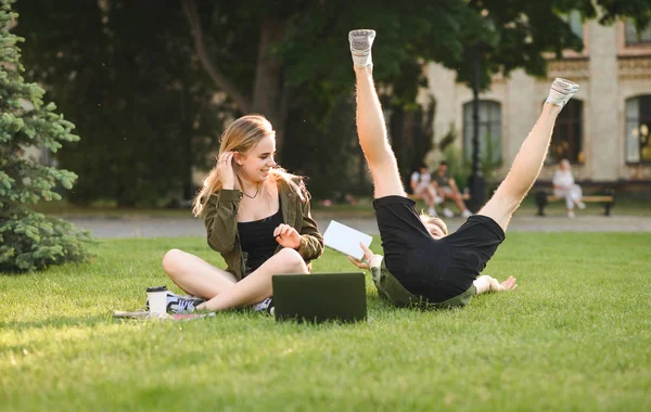 Caucasian cute couple of college students messing around on the lawn in the garden. Two funny students being silly, having fun in the university park, laptop, coffee and books near them.