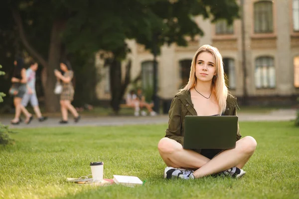 Στοχαστική μαθήτρια γυναίκα ονειροπόληση κοιτάζοντας πάνω ενώ δουλεύοντας σε ένα φορητό υπολογιστή που κάθεται στο Πανεπιστήμιο έξω σε πράσινα δέντρα γκαζόν και το Πανεπιστήμιο στο παρασκήνιο. Καυκάσιο κορίτσι με υπολογιστή, βιβλία και καφέ. — Φωτογραφία Αρχείου