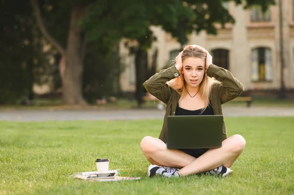 Aantrekkelijke jonge vrouwelijke student studeren hard buitenshuis op de campus zittend op het gazon met haar laptop met hoofdpijn het aanraken van haar hoofd, gevoel nerveus en gestrest. Overwerken, deadline concept. — Stockfoto