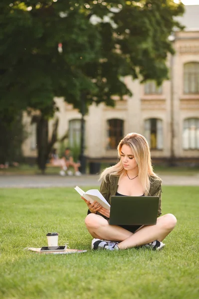 En positiv kvinnelig student som sitter med beina i kors på gress med laptop og bøker som gjør lekser og forbereder seg til timen. Konsentrert collegejente som sitter på plenen og leser bok. Vertikalt skudd . – stockfoto
