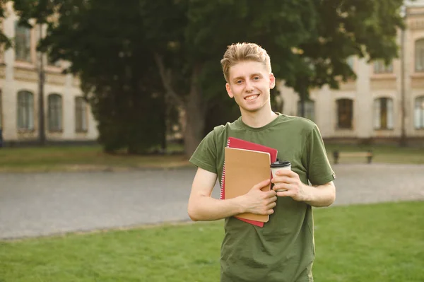 Handsome happy male college student standing in the park holding take away coffee and notebooks. Smiling positive university student boy near the campus with books and espresso cup.