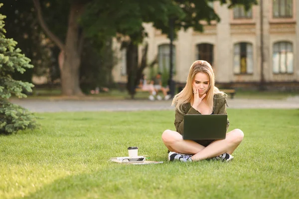 Zavarba fiatal női főiskolai hallgató olvasatban info rá laptop koncentrálódik, ül a gyepen. Egyetemi hallgató lány érzés zavaros nézi számítógép képernyőjén tartózkodó közelében Campus. — Stock Fotó