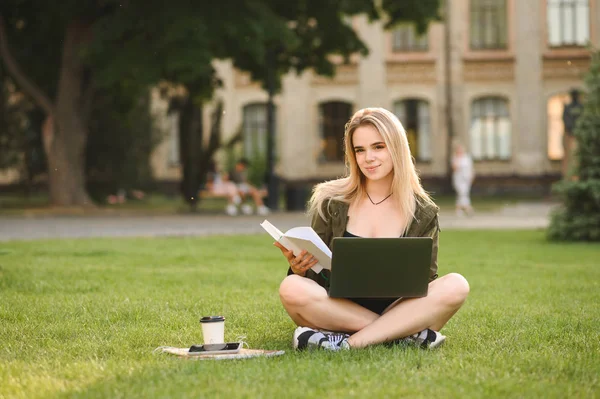 Positiv kvinnlig student sitter Cross-legged på gräs med laptop och böcker gör en läxa, tittar på kameran. Koncentrerad college flicka sitter på gräsmattan, läsa bok uppmärksamt. Horisontell bild. — Stockfoto