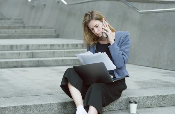 Портрет бізнес-леді, що сидить на вулиці на сходах, читаючи документи в її руках. Серйозна бізнес-леді, яка вивчає документи на вулиці на задньому плані міста з ноутбуком і кавою . — стокове фото