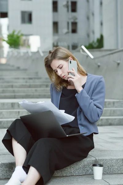 Портрет продуманого кавказького працівника жіночого офісу, що перевіряє документи, що розмовляють по телефону. Роботодавець сидить на сходах біля бізнес-центру з паперами, ноутбуком і розмовляє по телефону . — стокове фото