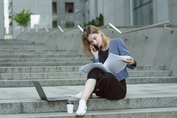 Joven empresaria concentrada leyendo los documentos sentada en las escaleras cerca del centro de negocios. Computadora portátil y café cerca del empleado diligente ocupado leyendo atentamente los periódicos . — Foto de Stock