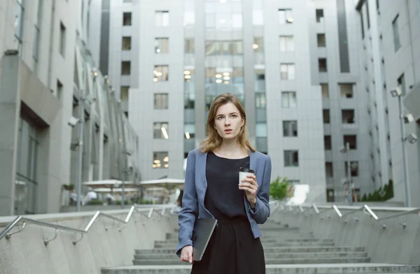 Pensive businesswoman walking around urban setting in financial district with coffee to go and laptop on concrete stairs. Thoughtful female office worker pondering holding her laptop and cup of coffee