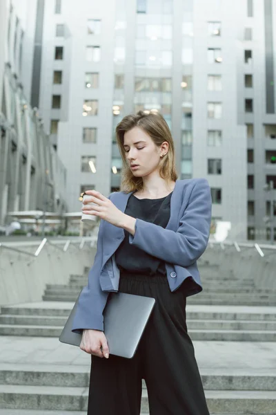 Beautiful businesswoman looking at coffee cup holding laptop standing on business center stairs. Vertical shot of female office worker at urban scenery with laptop, coffee in smart-casual clothes.