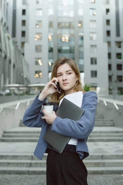 Feliz exitosa mujer de negocios llamando con teléfono celular cerca del edificio de oficinas sosteniendo laptop, documentos, café. Empresaria hablando por teléfono móvil cerca del centro de negocios mirando a un lado . — Foto de Stock