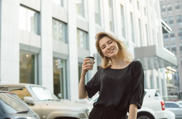 Χαμογελαστή ευτυχισμένη νεαρή γυναίκα κρατώντας ένα φλιτζάνι καφέ, φορώντας γκρι μπλούζα και μαύρο παντελόνι. Νεανικό χαρούμενο κορίτσι μπροστά από το αστικό τοπίο και χώρο στάθμευσης αυτοκινήτων. Πλατύ χαμόγελο. — Φωτογραφία Αρχείου