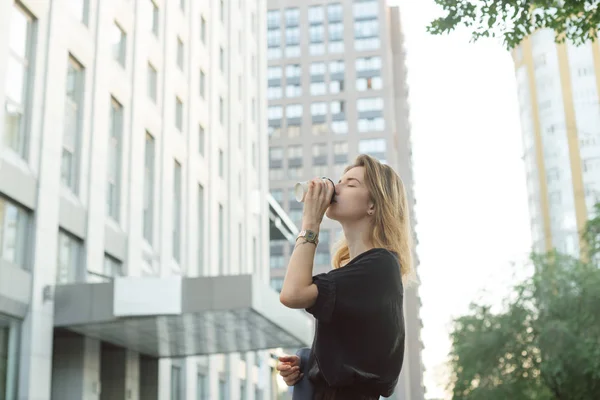 Весела стильна бізнес-леді п'є каву зовні на міському тлі. Приваблива молода жінка насолоджується кавою на вулиці біля офісної будівлі та хмарочоса . — стокове фото