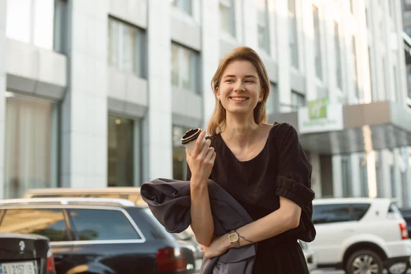 Vonzó kaukázusi lány nevetve és érzés boldog kívül a városi táj kávét iszik. Gyönyörű nő széles mosollyal gazdaságban elvenni kávé gyaloglás közelében az üzleti központ munka után. — Stock Fotó