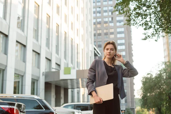 Foto de atractiva mujer de negocios en auriculares inalámbricos sosteniendo su computadora portátil mirando a la cámara cerca del edificio de oficinas. Empleado serio cerca del centro de negocios y estacionamiento en la calle con computadora . — Foto de Stock