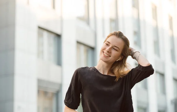 Retrato de menina sorridente em fones de ouvido sem fio perto do centro de negócios. Mulher atraente com amplo sorriso desfrutando de música nos fones de ouvido no distrito da cidade moderna . — Fotografia de Stock