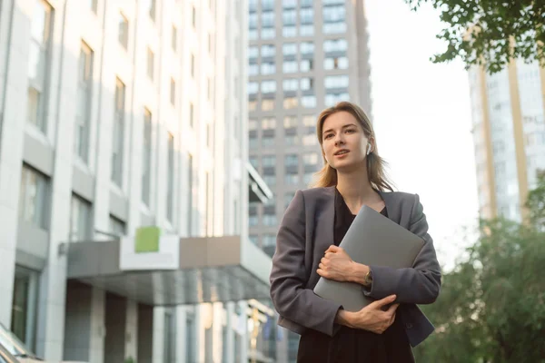 Portret van mooie zakenvrouw in Smart Casual kleding buiten het kantoorgebouw. Jonge vrouwelijke kantoormedewerker houdt laptop, glimlachend in haar draadloze koptelefoon in de buurt van het businesscentrum. — Stockfoto