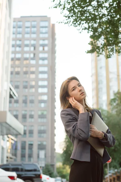 Joven empresaria exitosa en ropa casual inteligente frente a edificios de oficinas y rascacielos. Mujer sosteniendo un ordenador portátil moderno, escuchando música en auriculares inalámbricos en la calle al aire libre . — Foto de Stock