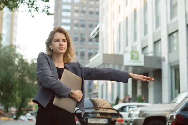 Joven profesional en un apuro enganchando un taxi en el distrito financiero, sintiéndose preocupada. Mujer de negocios concentrada tratando de coger un taxi cerca de los edificios de oficinas sosteniendo su computadora portátil . — Foto de Stock