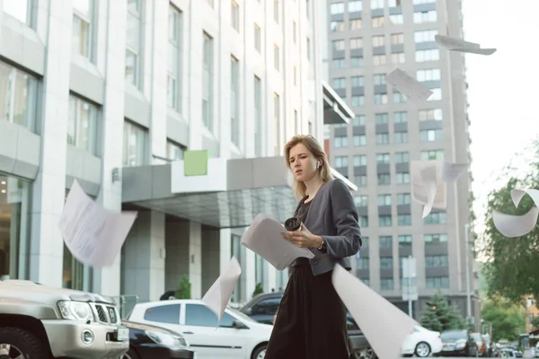 Una trabajadora de oficina confundida arrojando documentos al aire cerca del edificio de oficinas. Empresaria irritada lanzando papeles en el cielo sintiéndose desconcertada . — Foto de Stock