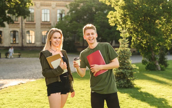 Dos amigos alegres, estudiantes con amplias sonrisas sosteniendo libros de texto y café en el parque cerca del edificio de la escuela secundaria. Pareja joven de estudiantes riendo cerca de la universidad con libros. Concepto educativo Fotos De Stock