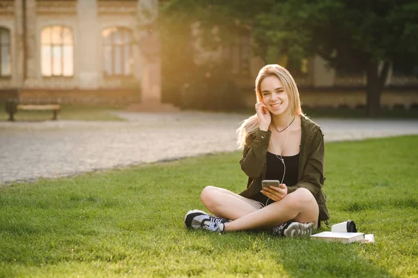 Estudiante sonriente escuchando música con un teléfono inteligente y auriculares sentados en el césped en un patio del campus. Chica alegre estudiante disfrutando de la música en el teléfono con auriculares cerca del edificio de la universidad . Imagen De Stock