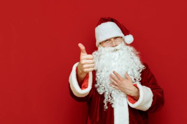 Klasik takım elbiseli ve sakallı pozitif Noel Baba kırmızı arka planda baş parmağını gösteriyor, kameraya bakıyor ve gülümsüyor, Noel Baba 'nın sevdiği gibi. Mutlu noel babanın portresi. Kopyalama. X mas konsepti