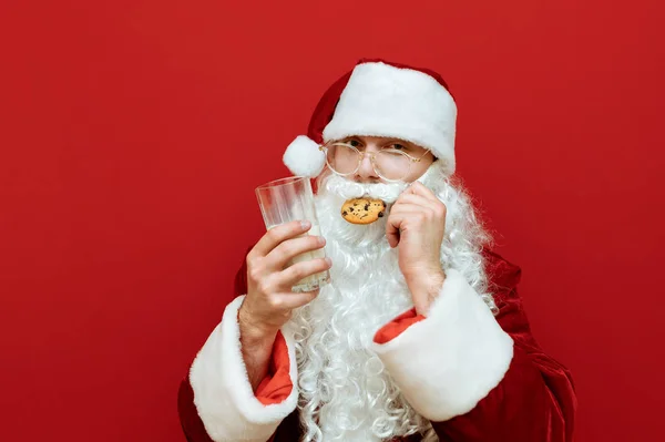Mężczyzna w kostiumie Świętego Mikołaja stoi na czerwonym tle ze szklanką mleka w ręku i je czekoladowe ciasteczko, patrząc w obiektyw z poważną twarzą. Mikołaj kocha świąteczne ciasteczka i mleko — Zdjęcie stockowe