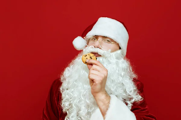 Portret pięknego Mikołaja z brodą i okularami trzymającego w ręku świąteczne ciasteczka i patrzącego w aparat z poważną twarzą. Tradycja z Mikołajem i ciasteczkami. Koncepcja świąteczna. — Zdjęcie stockowe